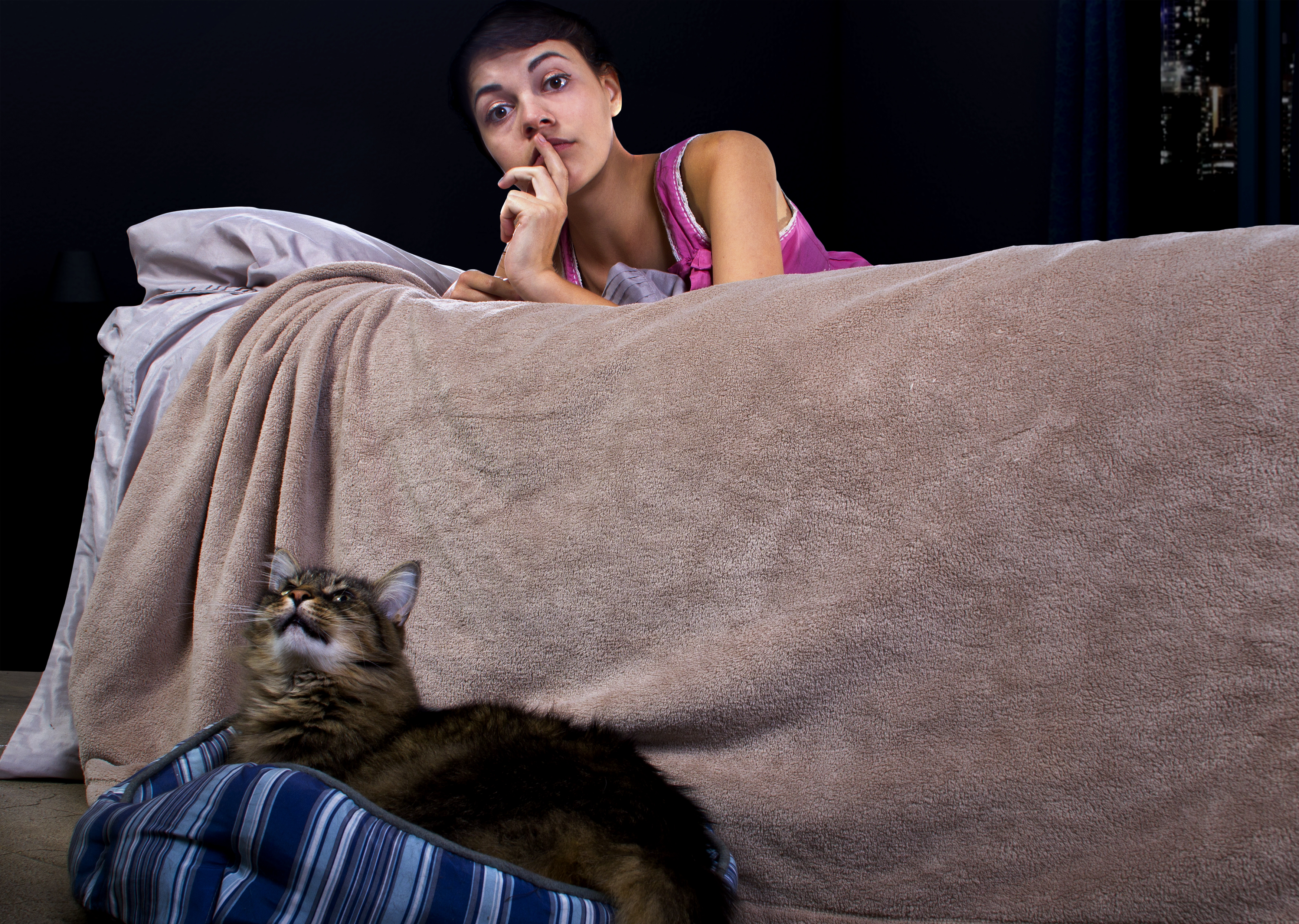 A sleepless woman in bed shushes a noisy cat sitting on the floor