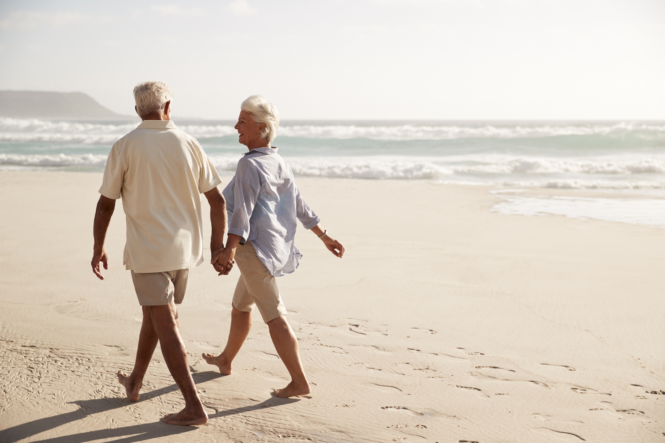 Happy older couple walks down a sunny beach holding hands